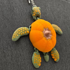 May include: A colorful turtle-shaped keychain with an orange body and green limbs, hanging on a gray fabric surface. The turtle has a detailed design and is attached to a silver chain.