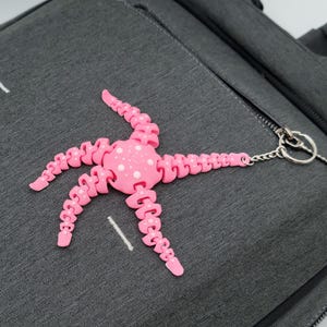 May include: A pink, articulated octopus charm with white polka dots is attached to a dark gray sling bag. The bag has a zippered pocket and a small metal charm on the zipper. White stitching accents the bag's surface.