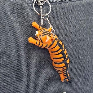 May include: A tiger-shaped keychain with orange and black stripes hangs from a silver keyring. The keychain is attached to a gray fabric surface, possibly a bag or backpack. The tiger is in a playful pose.