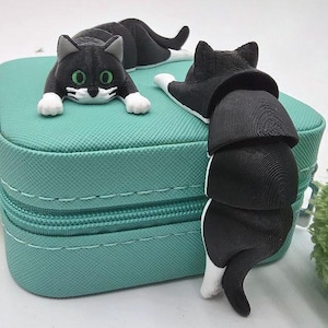 May include: A teal zippered case with two black and white cat figurines. One cat is lying down, and the other is climbing the side of the case. The cats have green eyes and white paws. An orange and green pipe cleaner is in the background.