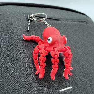 May include: A red octopus keychain with a white eye is attached to a gray bag. The octopus has articulated limbs and is made of plastic. The bag has a zipper and a small pocket.
