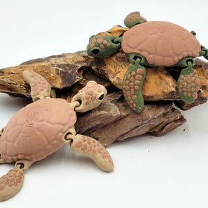 May include: Two toy turtles with articulated limbs and textured shells rest on a piece of brown, textured rock. The turtles have tan shells, green limbs, and tan patterned feet. The background is white.