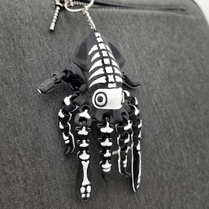 May include: A black and white squid keyring with a skeleton design hangs from a silver zip. The keyring is attached to a grey fabric surface. The squid has a detailed, stylized appearance.