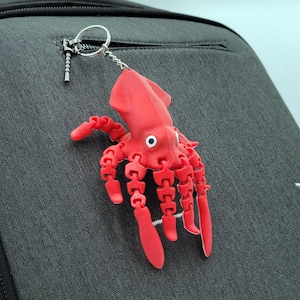 May include: A red articulated squid keychain with white eyes hangs from a silver keyring attached to a dark gray backpack. The squid's tentacles are segmented, and the backpack has a zippered pocket.