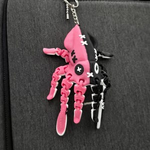 May include: A pink and black keychain in the shape of a stylized creature, suspended from a silver chain. The keychain is attached to a dark grey backpack. The creature has articulated limbs and a stitched design.