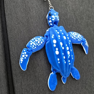 May include: A blue turtle-shaped keyring with white polka dots. The keyring is set against a dark grey fabric background. The turtle has articulated limbs and a silver metal ring for attachment.
