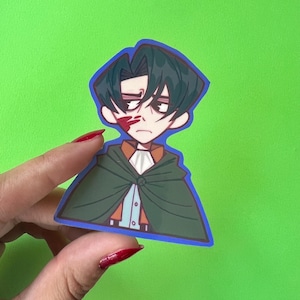 Puede incluir: Una pegatina de vinilo troquelada que muestra un personaje de anime estilizado con cabello oscuro, expresión seria y una marca roja en la mejilla. El personaje viste una capa verde oscuro sobre una camisa azul claro y una chaqueta marrón.