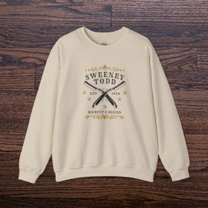 Op de afbeelding: Crèmekleurige sweatshirt met de tekst "Sweeney Todd" boven gekruiste rechte scheermessen. Het ontwerp bevat de tekst "EST. 1846" en "HAIRCUT & SHAVES" in vintage stijl. De sweatshirt heeft lange mouwen.