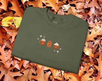 Può includere: Una maglietta verde con una grafica di tre bevande autunnali: un latte, una candela e un latte alla zucca. La grafica è in tonalità di marrone e arancione.