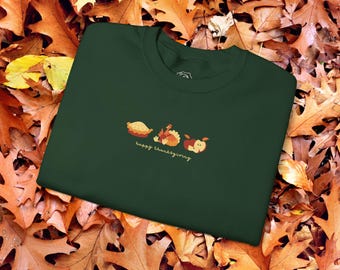 Può includere: Una maglietta verde scuro con un design a tema del Ringraziamento che presenta una torta, un tacchino e una mela. Il testo "happy thanksgiving" è stampato sotto il design.