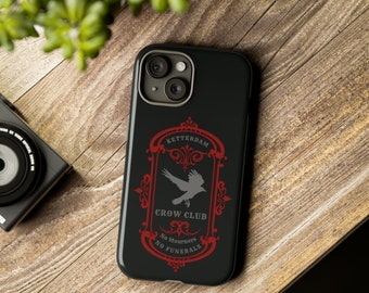 May include: Black phone case with a red and gray design. The design features the text "Ketterdam Crow Club" with a crow illustration. The case is on a wooden surface.