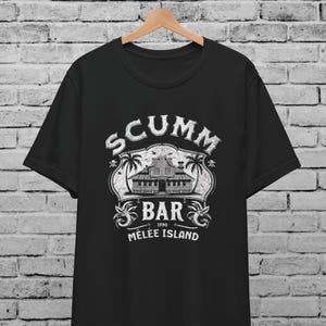 May include: Black t-shirt with a white graphic featuring the text "SCUMM BAR 1990 MELEE ISLAND". The graphic includes an illustration of a building, palm trees, and decorative elements. The shirt is hanging on a wooden hanger.