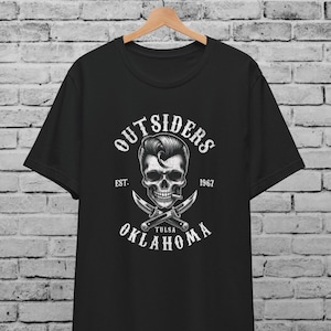 May include: Black t-shirt with a white graphic featuring a skull with a pompadour hairstyle, crossed knives, and the text "OUTSIDERS OKLAHOMA TULSA 1967". The shirt is hanging on a wooden hanger against a brick wall.