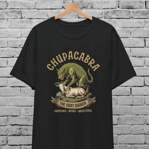 Pode incluir: Camiseta preta com uma ilustração verde e amarela de uma criatura mítica chamada Chupacabra atacando uma cabra. O texto "Chupacabra The Goat Sucker" está impresso na camiseta, juntamente com as localizações "Porto Rico, México, Estados Unidos".