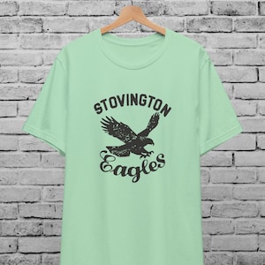 May include: A light green t-shirt with a black graphic of an eagle in flight. The graphic includes the text "Stovington Eagles".