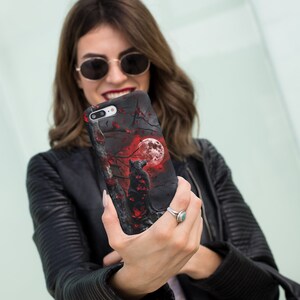 May include: A black phone case with a wolf howling at a red moon in a dark forest setting. The case has a red and black floral design.