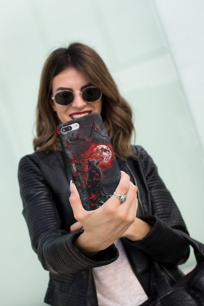 May include: A black phone case with a wolf howling at a red moon in a dark forest setting. The case has a red and black floral design.