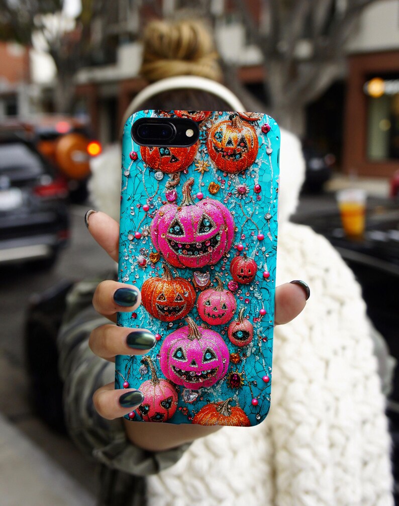 May include: A teal phone case featuring a Halloween-themed design. The case is adorned with orange and pink jack-o'-lanterns, sparkling gems, and decorative vines. The pumpkins have smiling faces and are of varying sizes, creating a festive and eye-catching accessory.