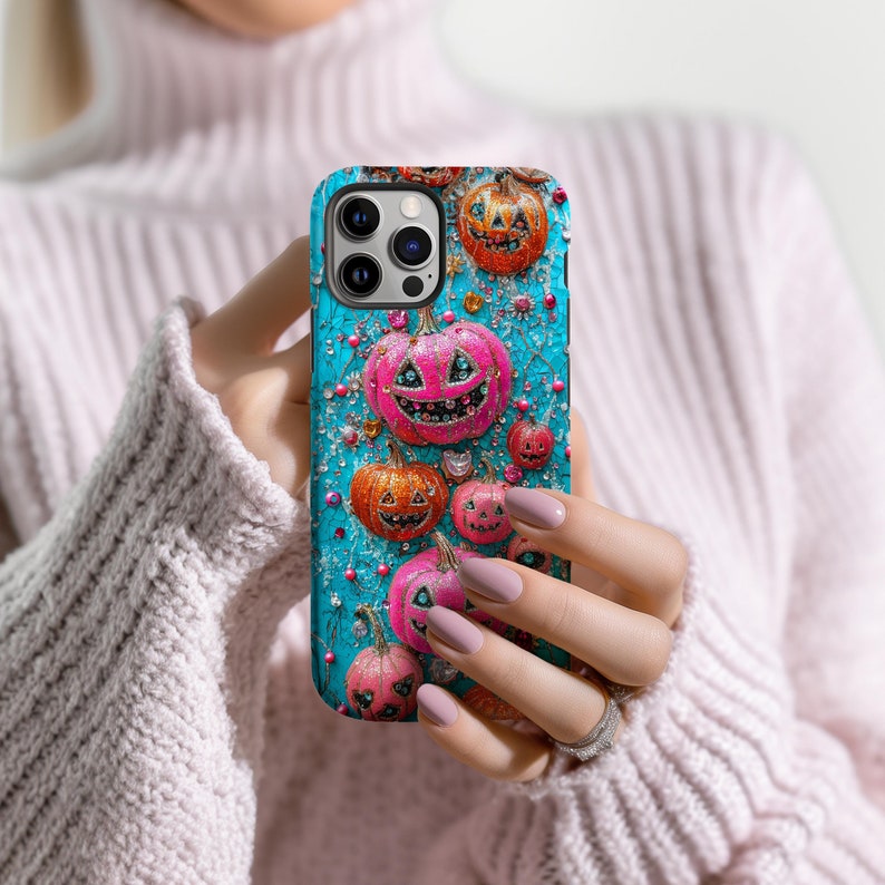 May include: A teal phone case featuring a Halloween-themed design. The case is adorned with orange and pink jack-o'-lanterns, with a textured background and sparkling accents. The case is held in a person's hands.