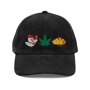 Op de afbeelding: Een zwarte corduroy baseballpet met drie geborduurde ontwerpen op de voorkant: een witte cartoonkip die een kippenpoot vasthoudt, een groen cannabisblad en een gele taart met een rasterkorst en rode kersen.