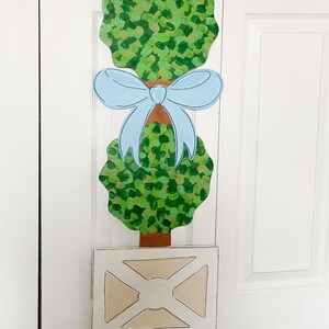 Peut inclure: Un arbre en bois en forme de topiaire avec deux buissons verts et un nœud bleu. L'arbre est dans un pot blanc avec une base brune et des roues.