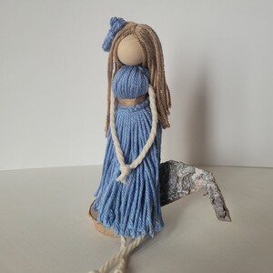 Puede incluir: Una muñeca de hilo azul con pelo de hilo marrón y un lazo de hilo blanco. La muñeca está sentada en un trozo de madera.