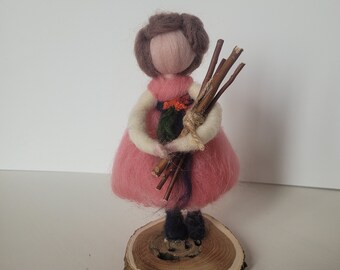 May include: A felt doll with brown hair, wearing a pink dress and holding a bundle of twigs. The doll is standing on a wooden base.