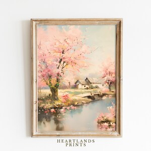 Puede incluir: Pintura de paisaje enmarcada que representa una escena serena con un río, un puente y edificios. Los árboles de flores rosas y los colores suaves crean una atmósfera pacífica. La obra de arte está en un marco de madera clara desgastada. El texto "HEARTLANDS PRINTS" está en la parte inferior.