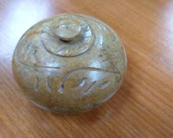 May include: A small, round, carved stone container with a lid. The container is a light brown color and has a decorative design carved into the surface.