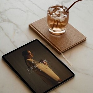 May include: A glass of iced beverage with a brown straw sits atop a brown textured notebook. A tablet displays an image with the text "TIFF'S LUX PE". The items are arranged on a marble surface.