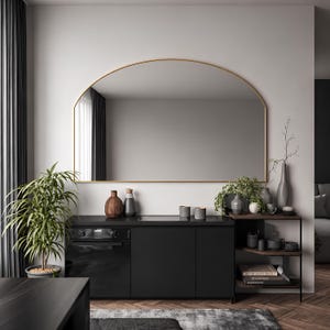 May include: A large arched wall mirror with a thin gold frame mounted above a black kitchen cabinet unit. The cabinet features a built-in oven, a smooth black countertop, and an adjacent open shelving unit holding various decorative ceramic vases.