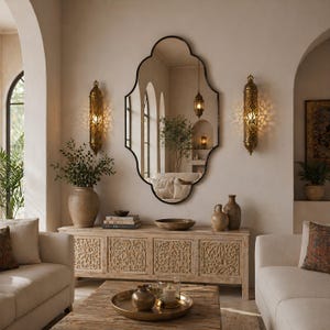 Peut inclure: Un grand miroir mural à bords festonnés doté d'un cadre noir fin, placé au-dessus d'une longue console en bois clair aux panneaux finement sculptés, encadré par deux appliques murales décoratives en métal doré diffusant une lumière.