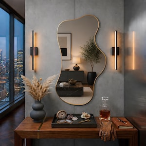 May include: An irregular, organic-shaped wall mirror with a thin gold frame. The mirror reflects a room interior featuring a potted plant, a table lamp, and a sideboard. It is mounted on a gray concrete wall between two vertical black wall sconces.