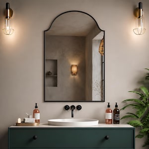 May include: An arched wall mirror with a thin black frame positioned above a white oval vessel sink and a dark green vanity cabinet. The mirror reflects a room with a wall sconce, a recessed shelf, and a woven light fixture hanging nearby.