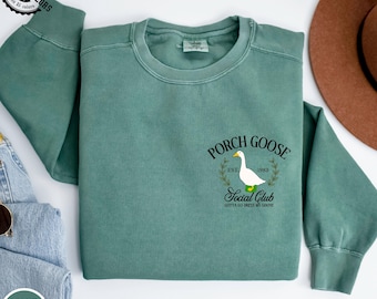 Könnte beinhalten: Ein hellgrünes Sweatshirt mit dem Text "PORCH GOOSE Social Club" und einer Gänsegrafik. Das Sweatshirt besteht aus 100 % Baumwolle und trägt die Aufschrift "Comfort Colors". Der Text "GOTTA GO DRESS MY GOOSE" ist ebenfalls auf dem Sweatshirt aufgedruckt.