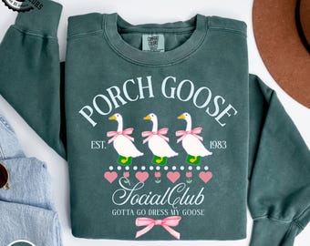 Könnte beinhalten: Blau-Fichte-Sweatshirt mit dem Text "PORCH GOOSE" und "Social Club". Die Grafik zeigt drei Gänse mit rosa Schleifen, rosa Herzen und Tulpen. Der Text "GOTTA GO DRESS MY GOOSE" ist ebenfalls enthalten.