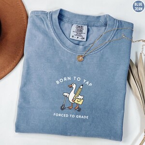 May include: A blue jean Comfort Colors t-shirt with the text "BORN TO YAP" and "FORCED TO GRADE". The graphic features a cartoon goose riding a scooter, holding a pencil and a briefcase. A gold necklace is draped across the shirt.