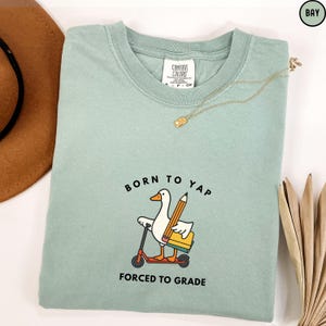May include: A light green t-shirt with the text "BORN TO YAP FORCED TO GRADE" and a cartoon duck riding a scooter while holding a pencil and a book. The shirt has a crew neck and short sleeves.