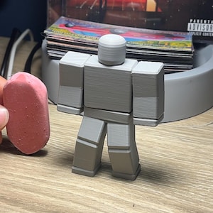 May include: A gray, 3D-printed figure with a blocky design, resembling a popular video game character. The figure has a cylindrical head and rectangular body parts. A pink eraser is next to the figure.