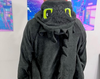May include: Black hooded sweatshirt with a dragon design. The hood features large, bright green eyes and a detailed face. The back of the sweatshirt has a row of stylized spines. The sweatshirt is made of a soft, plush material.
