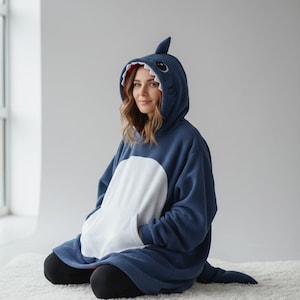 May include: A dark blue oversized fleece hoodie designed to resemble a shark. The hood features a shark face with white teeth and a dorsal fin on top. The front includes a large white circular panel and a kangaroo pocket, with a tail at the back.