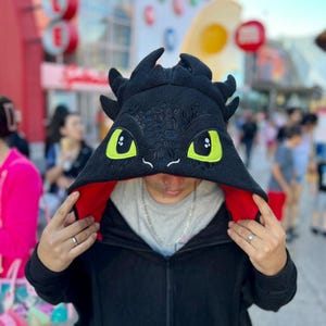 May include: Black hooded sweatshirt featuring a dragon design. The hood is black with embroidered details and large yellow eyes. The interior of the hood is red. The sweatshirt has a zipper closure and a dragon tail design on the back.