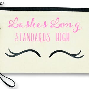 Könnte beinhalten: Eine weiße Canvas-Schminktasche mit einem schwarzen Reißverschluss und einem schwarzen Riemen. Die Tasche hat einen pinkfarbenen Text mit der Aufschrift "Lashes Long Standards High" und eine schwarze Illustration von Wimpern.