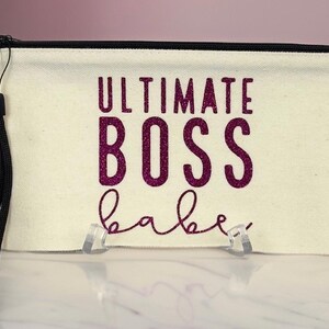 Puede incluir: Una bolsa de lona blanca con cremallera y cierre de cremallera negro y una correa negra. La bolsa tiene letras de purpurina rosa que dicen "ULTIMATE BOSS babe".