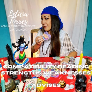 May include: A woman wearing a blue bandana and a white mesh top sits at a table with a red tablecloth. She is holding a heart-shaped card with a picture of a couple on it. The table is decorated with candles, tarot cards, and other spiritual items. The text on the table reads "COMPATIBILITY READING STRENGTHS WEAKNESSES ADVISES".