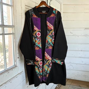 Peut inclure: Un cardigan noir à manches longues doté d'un col côtelé et d'un panneau central vertical avec un motif floral et géométrique coloré dans des tons de violet, bleu sarcelle, rouge et blanc. Le vêtement porte une étiquette de marque Koos au col.