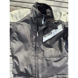 Puede incluir: Chaqueta de moto negra con cuello alto y cierre de cremallera. La chaqueta tiene hombreras grises y un parche blanco y plateado con la inscripción "ROAD CRAFTER". Tiene dos bolsillos delanteros.