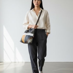 Puede incluir: Una mujer con una camisa crema y pantalones plisados grises oscuros lleva un bolso de hombro. El bolso presenta un diseño de patchwork en tonos grises, blanco, amarillo y marrón. Tiene una correa marrón oscuro.