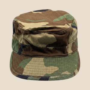 May include: A military-style patrol cap featuring a woodland camouflage pattern in shades of olive green, brown, tan, and black. The hat has a flat, circular top, a structured vertical band, and a short, curved brim with reinforced stitching.