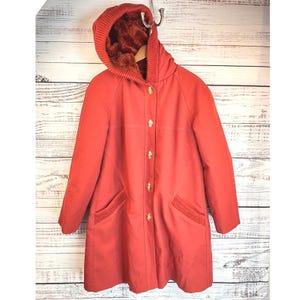 May include: A vintage red coat with a hood lined with brown faux fur. The coat has gold-coloured buttons and ribbed knit detailing on the pockets. The coat is hanging on a metal hook against a wooden background.